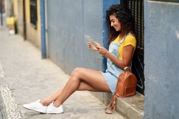 Young Arab woman looking at her digital tablet outdoors. African girl wearing casual clothes inurban background. Curly hairstyle.. Young Arab woman looking at her digital tablet outdoors.