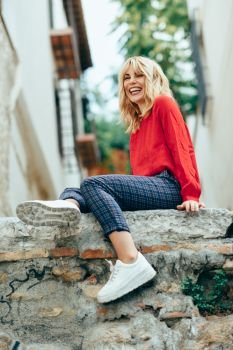 Happy young blond woman sitting on urban background. Smiling blonde girl with red shirt enjoying life outdoors.. Smiling blonde girl with red shirt enjoying life outdoors.