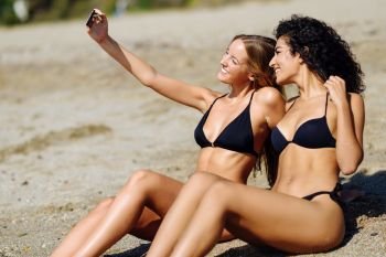 Two women taking selfie photograph with smartphone in the beach, sittingo on the sand.. Two women taking selfie photograph with smartphone in the beach