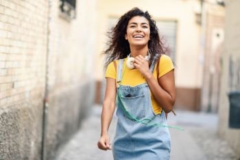 Young African woman with headphones and black curly hairstyle walking outdoors. Happy girl wearing yellow t-shirt and denim dress in urban background.. Young African woman with headphones walking outdoors