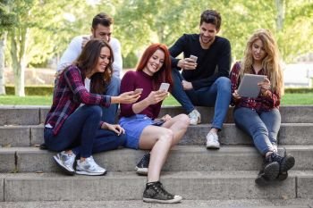 Group of young people using smartphone and tablet computers outdoors in urban background. Women and men sitting on stairs in the street wearing casual clothes.. Young people using smartphone and tablet computers outdoors