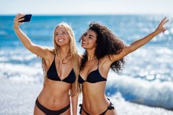 Two young women taking selfie photograph with smart phone in swimwear on a tropical beach. Funny caucasian and arabic females wearing black bikini.. Two women taking selfie photograph with smartphone in the beach