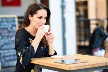 Middle-aged woman drinking coffee in an urban cafe bar. Young female sitting at table at an outside terrace. Girl with california highlights hairstyle.. Middle-aged woman drinking coffee in an urban cafe bar.