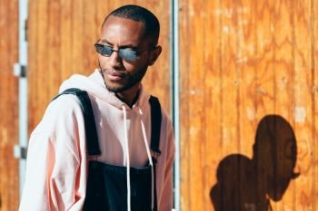 Millennial black man wearing casual clothes and sunglasses, against a wooden background. African guy with bib pants outdoors. Millennial African guy with bib pants outdoors