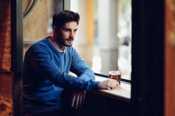 Young man with blue sweater with lost look in a modern pub. Thoughtful guy with modern hairstyle near a window drinking a soda.. Pensive guy with modern hairstyle near a window drinking a soda.