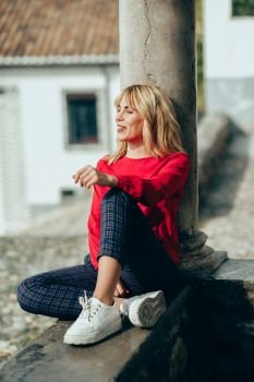 Happy young blond woman sitting on urban background sunbathing in autumn. Smiling blonde girl with red shirt enjoying life outdoors.. Happy young woman sitting on urban background sunbathing in autumn