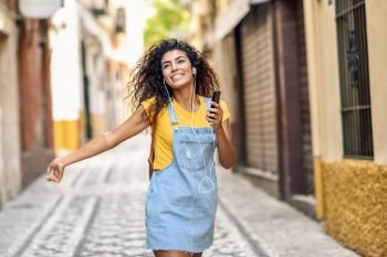 Attractive African woman listening to music with earphones outdoors. Black girl in casual clothes with curly hairstyle in urban background.. Young black woman listening to music with earphones outdoors