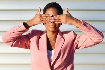 Funny black woman, model of fashion, standing on urban wall covering her eyes with her hands. African american female wearing suit with pink jacket with sunset light.. Funny black woman standing on urban wall covering her eyes with her hands.