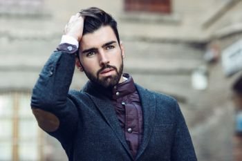 Young bearded man, model of fashion, in urban background wearing british elegant suit. Guy with beard and modern hairstyle touching his hair. Young bearded man wearing british elegant suit touching his hair