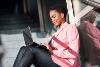 Smiling black businesswoman sitting on urban steps working with a laptop computer. African american female wearing suit with pink jacket.. Smiling black businesswoman sitting on urban steps working with a laptop computer