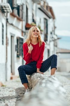Happy young blond woman sitting on urban background. Laughing blonde girl with red shirt enjoying life outdoors.. Smiling blonde girl with red shirt enjoying life outdoors.