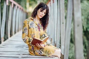 Young woman, wearing flowered dress, reading a book on a rural bridge.. Young woman reading a book on a rural bridge.