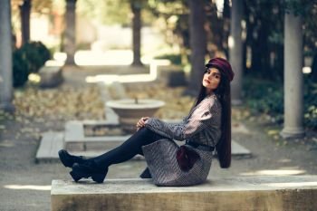Young beautiful girl wearing winter coat and cap sitting on a bench in urban park. Lifestyle and fashion concept.. Young beautiful girl wearing winter coat and cap sitting on a bench in urban park.