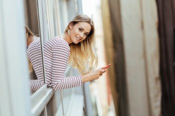 Young blonde woman leaning out of her house window using a smart phone.. Young woman leaning out of her house window using a smartphone.