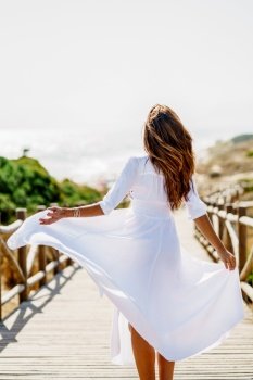 Rear view of a young woman wearing a beautiful white dress in Spanish fashion on a boardwalk on the beach.. Young woman wearing a beautiful white dress in Spanish fashion on a boardwalk on the beach.