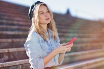 Blonde woman using her smartphone sitting on some city steps, wearing a denim shirt and black beret.. Blonde woman using a smartphone sitting on some city steps, wearing a denim shirt and black beret.