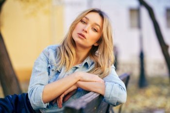 Blonde woman wearing denim shirt and black leather skirt sitting in an urban bench. Pretty russian female.. Blonde woman wearing denim shirt and black leather skirt sitting in an urban bench.