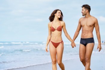 Young couple of beautiful athletic bodies walking together hand by hand on the beach enjoying their holiday at sea. Young couple of beautiful athletic bodies walking together hand by hand on the beach