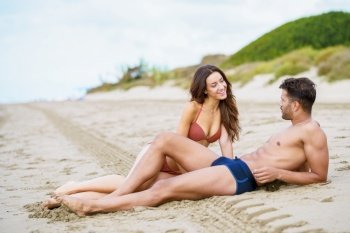 Young couple of beautiful athletic bodies sitting together on the sand of the beach enjoying their holiday at sea. Young couple sitting together on the sand of the beach