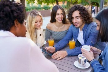 Young man showing something to his multi-ethnic group of friends on his smartphone, while having drinks at an outdoor table in a bar.. Guy showing his smartphone to his group of friends while having drinks at an outdoor bar