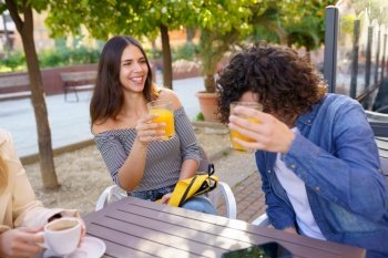 Couple of friends toasting with orange juices while having a drink with their multi-ethnic group of friends at the outdoor table of a bar.. Couple of friends toasting while having a drink with their multi-ethnic group of friends