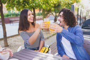 Couple of friends toasting with orange juices while having a drink with their multi-ethnic group of friends at the outdoor table of a bar.. Couple of friends toasting while having a drink with their multi-ethnic group of friends