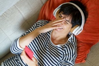 Woman laughing looking at her smartphone while listening to music with headphones lying on the sofa.. Woman laughing looking at her smartphone while listening to music with headphones.