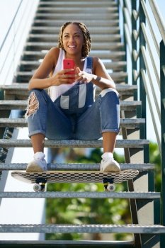 Black girl with coloured braids, sitting on some steps with a smartphone and a skateboard.. Black girl with coloured braids, sitting with a smartphone and a skateboard.