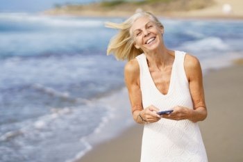Smiling mature woman walking on the beach using a smartphone. Elderly female enjoying her retirement at a seaside location.. Smiling mature woman walking on the beach using a smartphone.