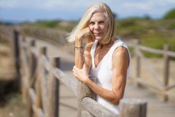Woman in her 60s leaning on wooden fences on the sand of a tropical beach, enjoying her time of relaxation. Mature female looking at camera.. Happy female in her 60s leaning on wooden fences on the sand of a tropical beach.
