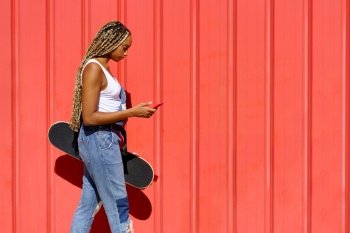 Young black girl with coloured braids walking on an urban red background while consulting her smartphone.. Black girl walking on an urban red background while consulting her smartphone.