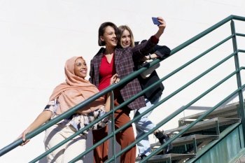 From below of positive young diverse female best friends smiling happily while taking selfie on smartphone standing on metal staircase in city on sunny day. Cheerful multiracial girlfriends taking selfie on smartphone on stairs