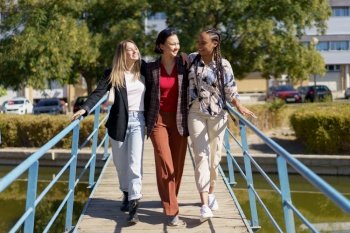 Full body of cheerful multiracial girlfriends in stylish clothes walking on footbridge over canal and looking at each other. Smiling diverse friends hugging while strolling on narrow footbridge