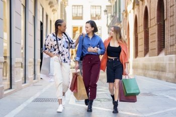 Full body of happy young trendy diverse female friends in stylish outfits smiling while walking together on city street with paper bags after successful shopping. Stylish multiracial female shopaholics walking together on street with paper bags