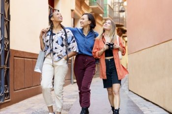 Cheerful young multiethnic female travelers in stylish clothes smiling and looking up while sightseeing old town during walk on street with photo camera. Happy multiracial female best friends walking in city during holidays