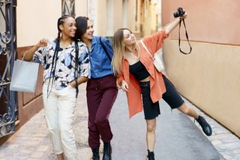 Content young multiethnic female best friends in stylish outfits smiling and taking selfie on photo camera while walking in old town during weekend. Happy diverse ladies taking selfie on camera during trip in city