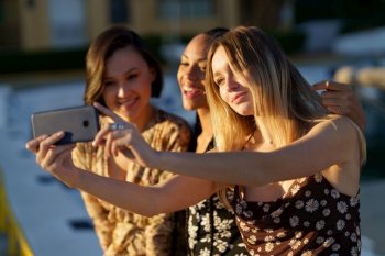 Woman taking self portrait on cellphone with positive young multiethnic girlfriends while enjoying time together. Charming diverse girlfriends taking selfie on smartphone