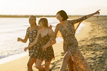 Happy multiethnic girlfriends in summer dresses holding hands and hugging while walking on sandy beach near sea in sunny evening. Laughing diverse women enjoying sunset on beach