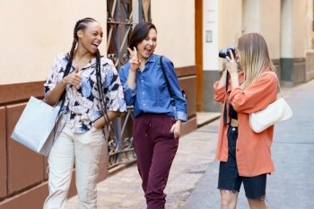Joyful young multiracial female tourists, smiling and winking while showing v sign and thumb up gesture, during walk on city street with female friend taking picture on photo camera. Content lady taking photo of delighted female friends on street