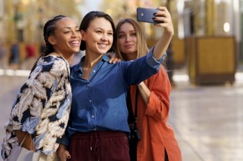 Confident young multiethnic female friends in stylish clothes smiling and taking self portrait on smartphone standing on city street during holidays. Content young multiracial female tourists smiling at taking selfie on smartphone