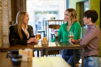 Happy adult women with alcohol drinks smiling and talking while gathering around table during friends meeting in pub. Cheerful girlfriends gathering around table in bar