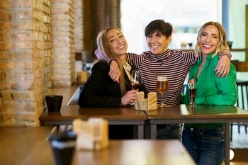 Cheerful group of female friends looking at camera with smile and embracing girlfriends behind table with alcohol during meeting in pub. Cheerful girlfriends hugging during party in bar