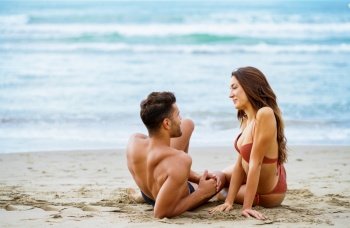 Cheerful loving woman in bikini and man in shorts sitting on shore in summer and looking at each other. Caucasian couple on sandy beach near sea