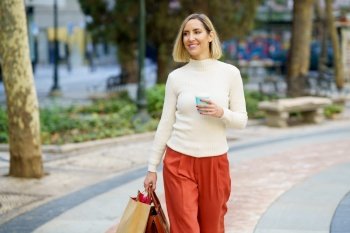 Positive female walking with paper bag and handbag and holding cup of takeaway beverage in park and looking away. Smiling woman with takeaway coffee carrying paper bag