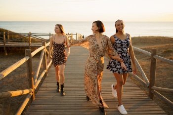 Full body of positive multiracial female friends strolling on wooden footbridge while spending time on seashore near sea on sunny day. Cheerful diverse girlfriends walking on footbridge near sea