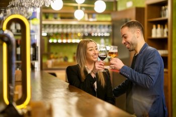 Happy diverse girlfriend and boyfriend looking at each other with smile and clinking glasses of alcohol drinks near counter during date in bar. Multiethnic couple proposing toast in a pub
