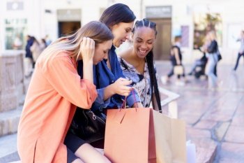 Satisfied diverse women sitting on street with paper bags and looking down while discussing purchases. Multiracial females spending time together on street after shopping