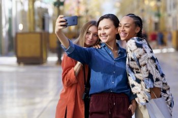 Happy diverse female friends taking self portrait on smartphone while standing close to each other on walkway in city against blurred background. Smiling multiracial women taking selfie in city