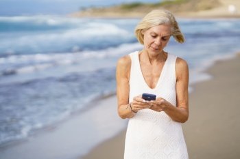 Senior woman walking on the beach using a smartphone. Elderly female enjoying her retirement at a seaside location.. Senior woman walking on the beach using a smartphone.
