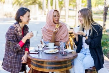 Smiling multiracial female friends eating and drinking coffee while sitting at table on terrace of outdoor cafeteria on summer day. Laughing diverse women in outdoor cafe